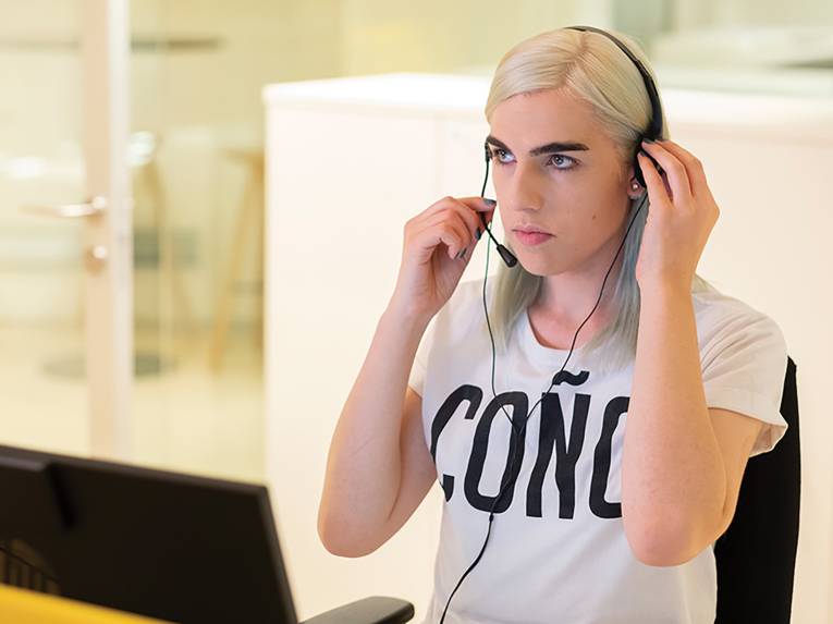 Das Bild zeigt eine blonde Person mit markanten, dunklen Augenbrauen, die das Headset auf ihrem Kopf gerade rückt. Die Person sitzt in einem Bürostuhl und trägt ein weißes Tshirt mit schwarzer Aufschrift.